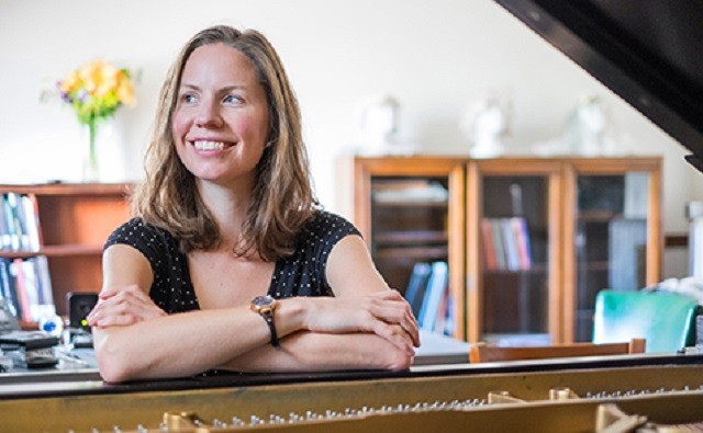 Kate Soper smiling at a piano and looking to the viewer's left. There are flowers and busts of statues in the out-of-focus background.