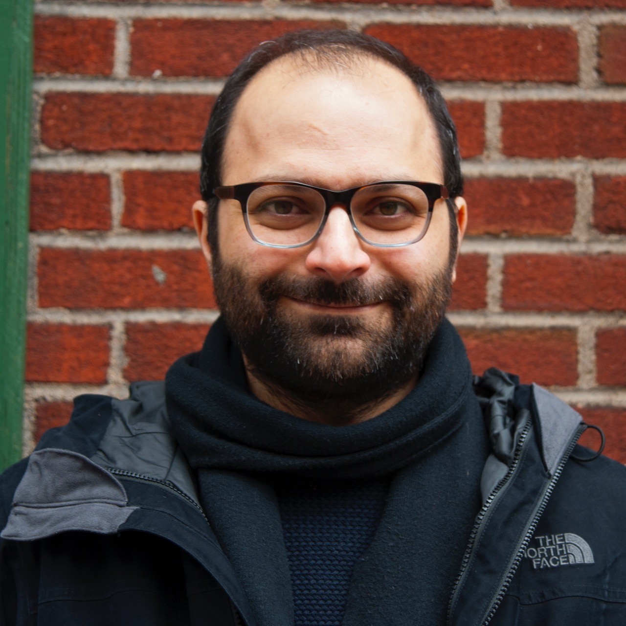 Ashkan Behzadi, in a dark blue sweater, scarf and jacket, stands in front of a red brick background. He is smiling at the camera.