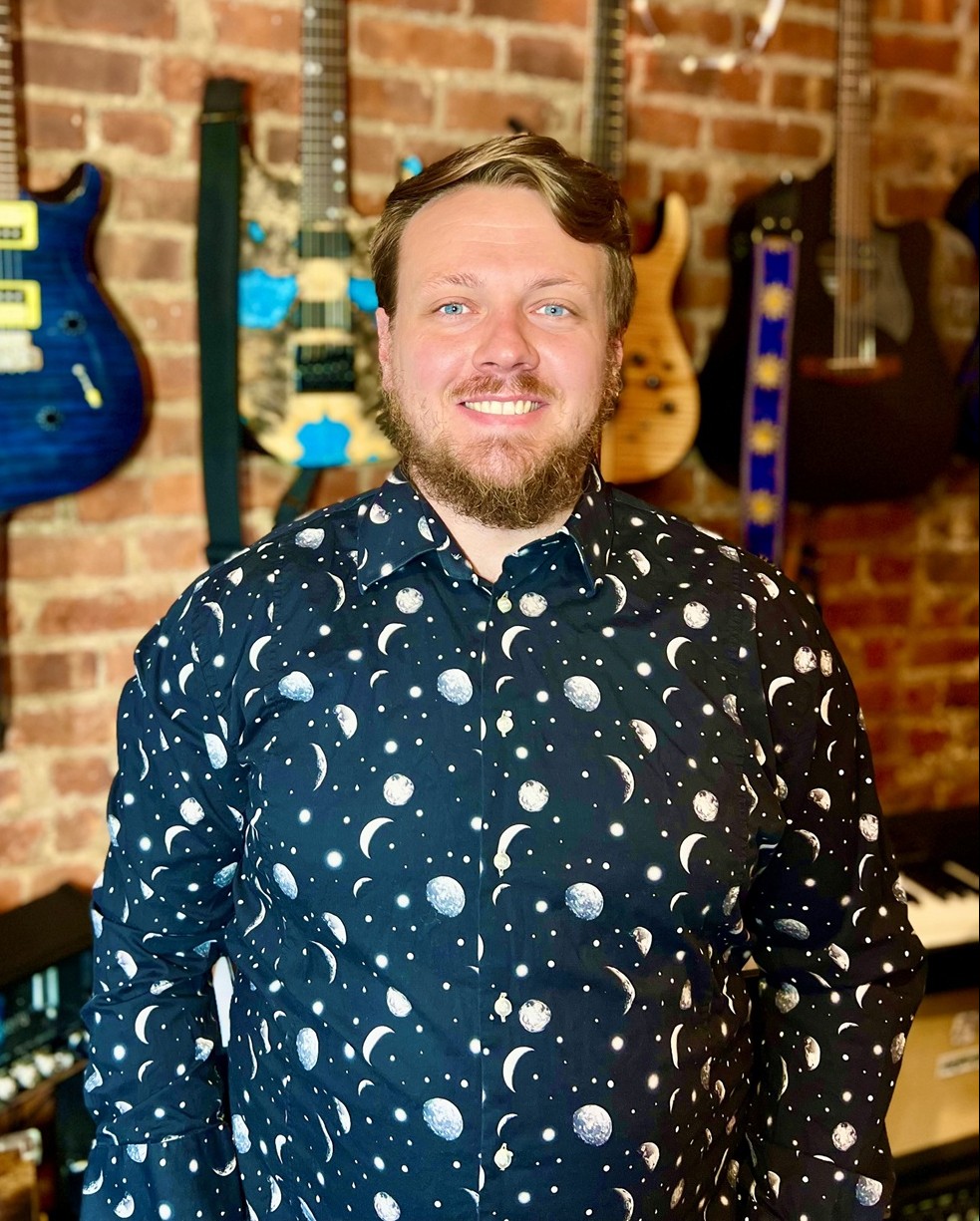 Calder Hannan standing in front of a brick wall with many guitars hanging from it.