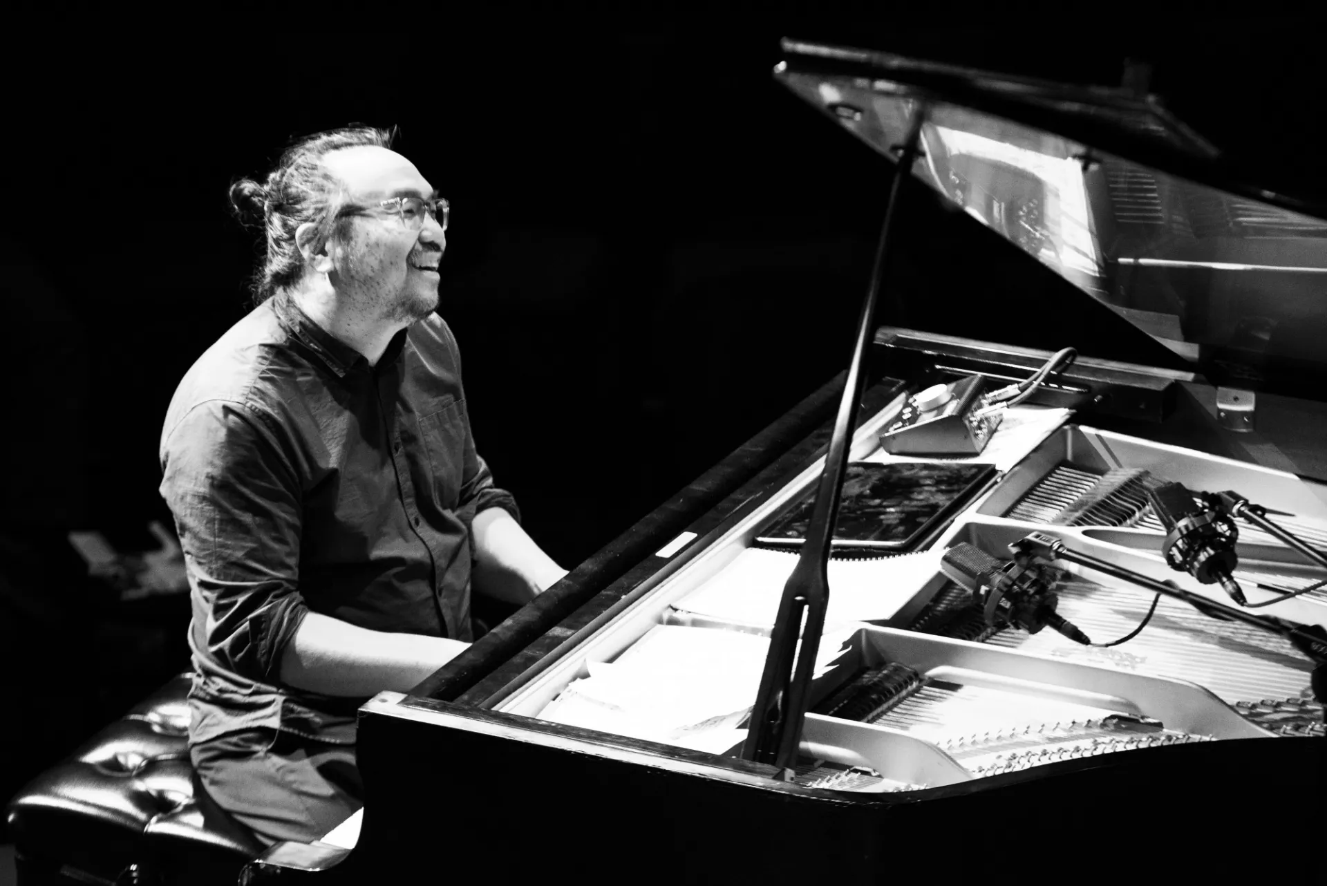 Black and white photo of John Chin at a piano. He is to the left of the image and is smiling toward the right side of the photo.