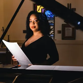 Courtney Bryan in a black dress at a piano holding sheet music
