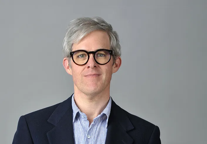 Ryan Dohoney in front of a gray background. He has gray hair, black glasses, and a light blue dress shirt under a dark blue coat.
