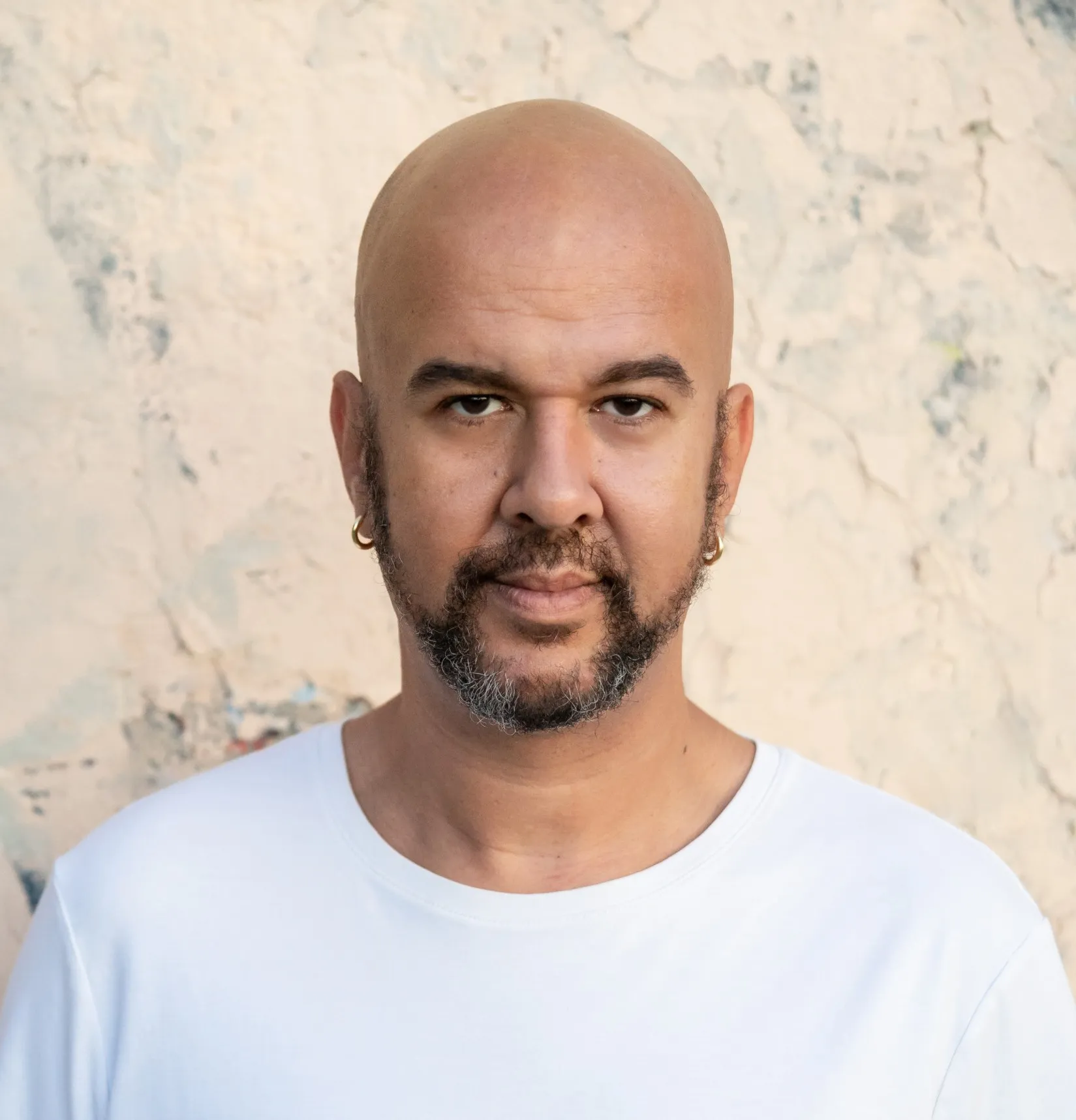 Marcos Balter in a white shirt in front of a pale background. He is bald, with a beard and two gold earrings.