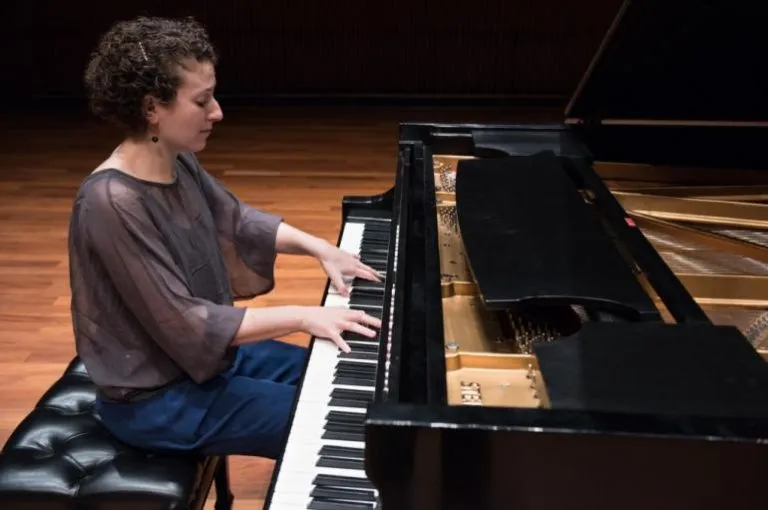 Pianist Clare Longendyke at a grand piano. She has curly brown hair, blue jeans, and a sheer gray top.