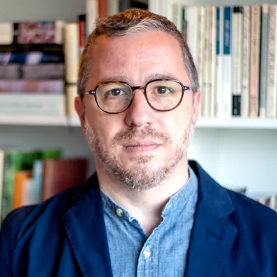 Seth Cluett in a light blue button up and dark blue jacket. He is looking straight at the camera with a wall of books out of focus behind him. He has calico glasses and a speckled gray beard, and he is wearing a neutral expression on his face.