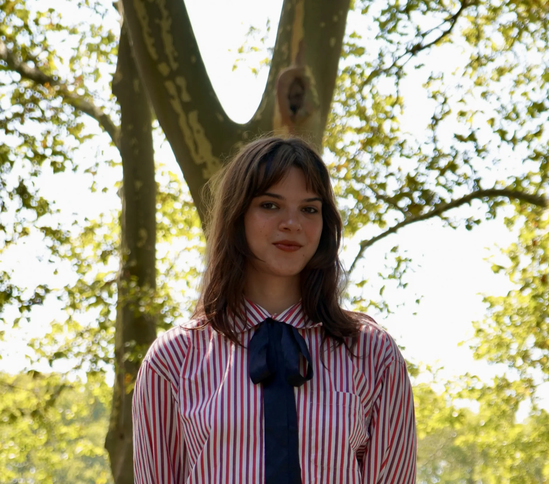 Azalea in a pink button down and dark blue scarf/tie hybrid. She stands in front of a tree with a slight smile.
