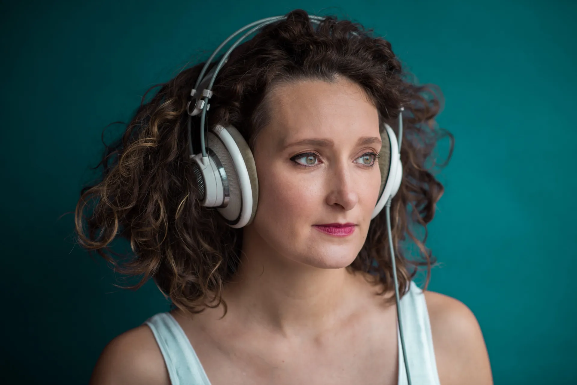 Zosha Di Castri in a white top wearing silver headphones. She is looking to the right of the image, and she is in front of a deep teal background.