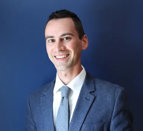 Scott Douglass in a blue suit in front of a blue background. He's smiling at the camera.