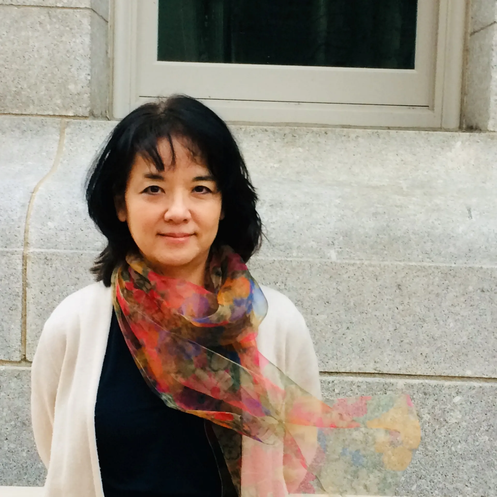 Ellie Hisama with a multi-colored scarf, white sweater and black shirt in front of light granite stonework.
