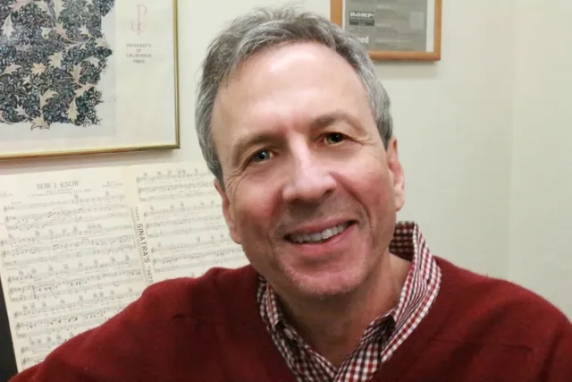 Walter Frisch smiling at the camera. He is dressed in red, with a checkered shirt under a crimson sweater.