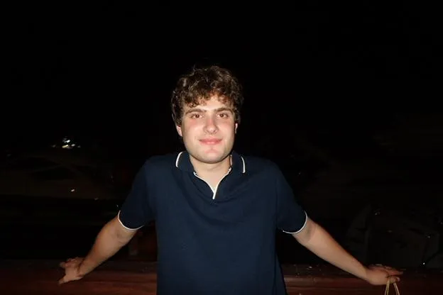 Max Kleban smiling at the camera against a very dark background. He's wearing a dark blue polo shirt.