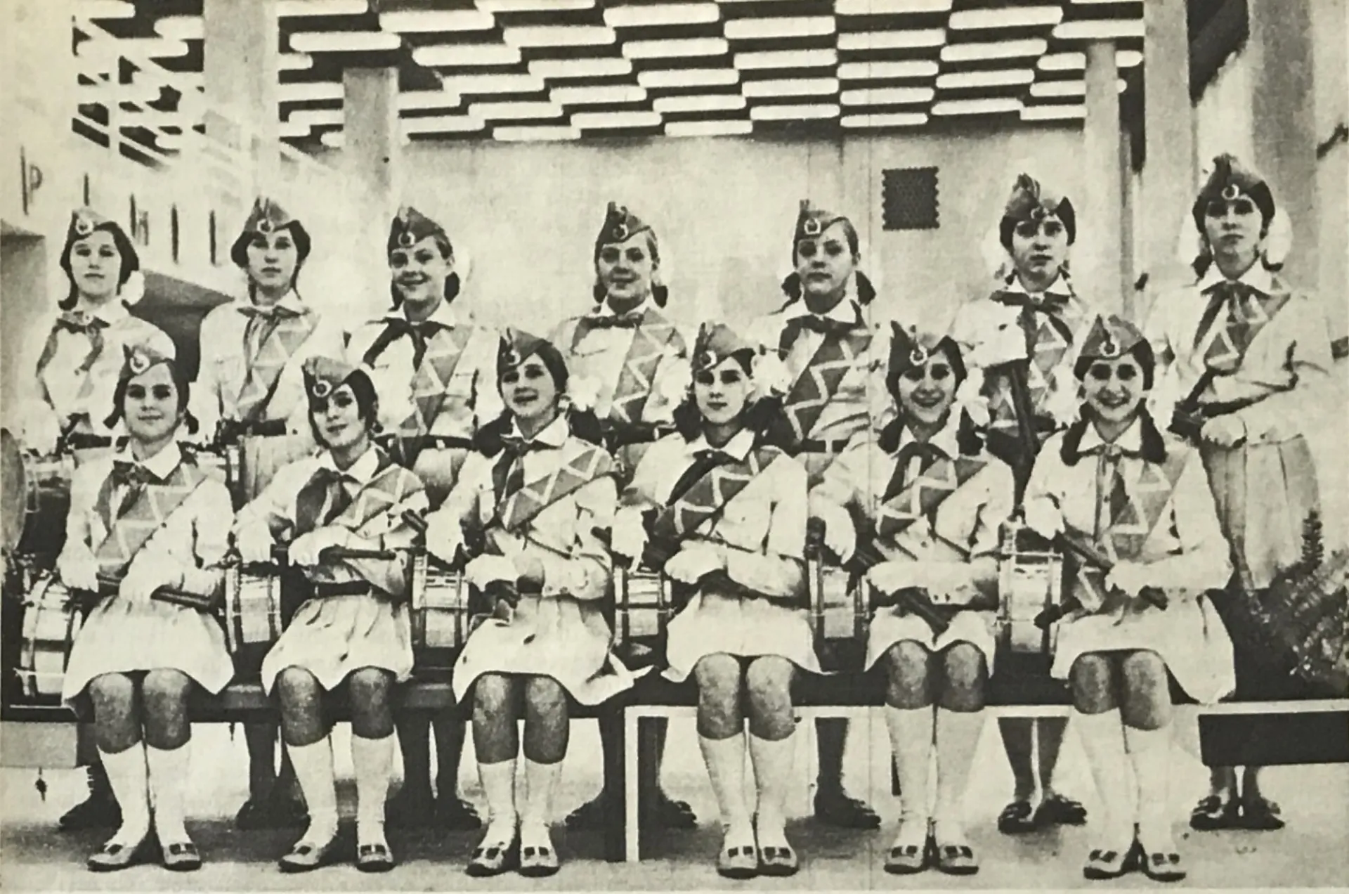 Attribution to the KPDU Archive. 1973 Palace of Pioneers drumming ensemble. Black and white photo of 13 girls in matching dresses, hats and sashes. They are sitting and standing and holding drums at their sides.