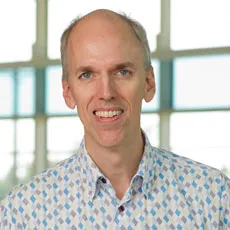 Michael Klein in a white and blue patterned shirt, smiling in front of windows.