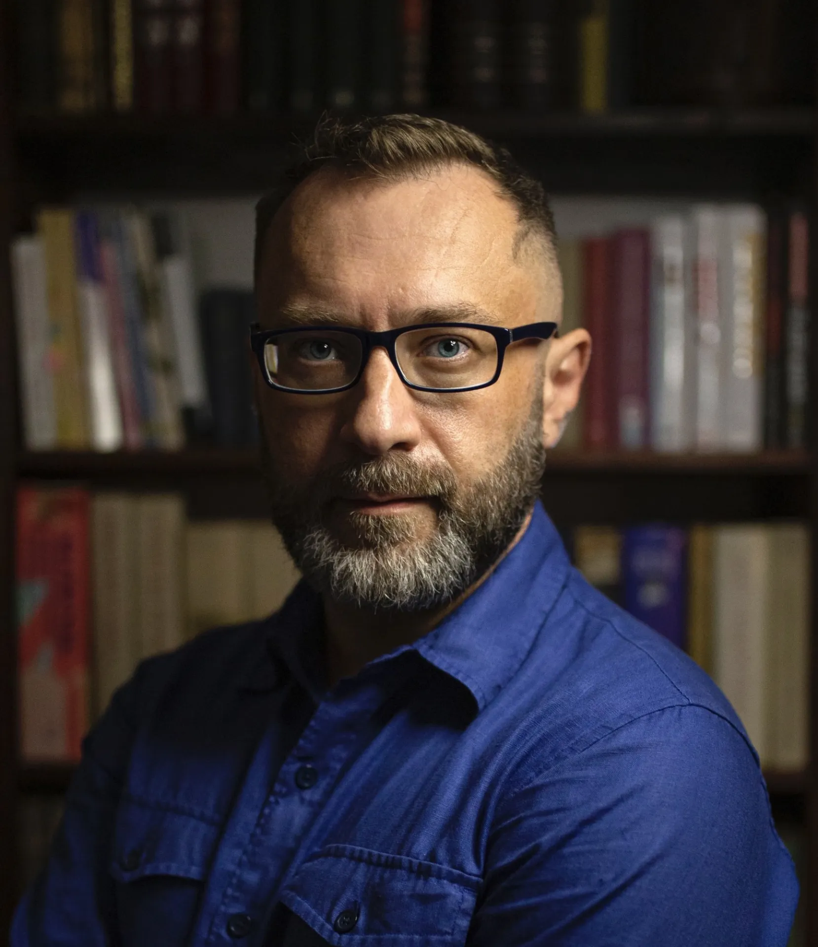 Photo of Mariusz Kozak in front of a dark bookshelf brimming with books. He is wearing glasses and a blue collared shirt.