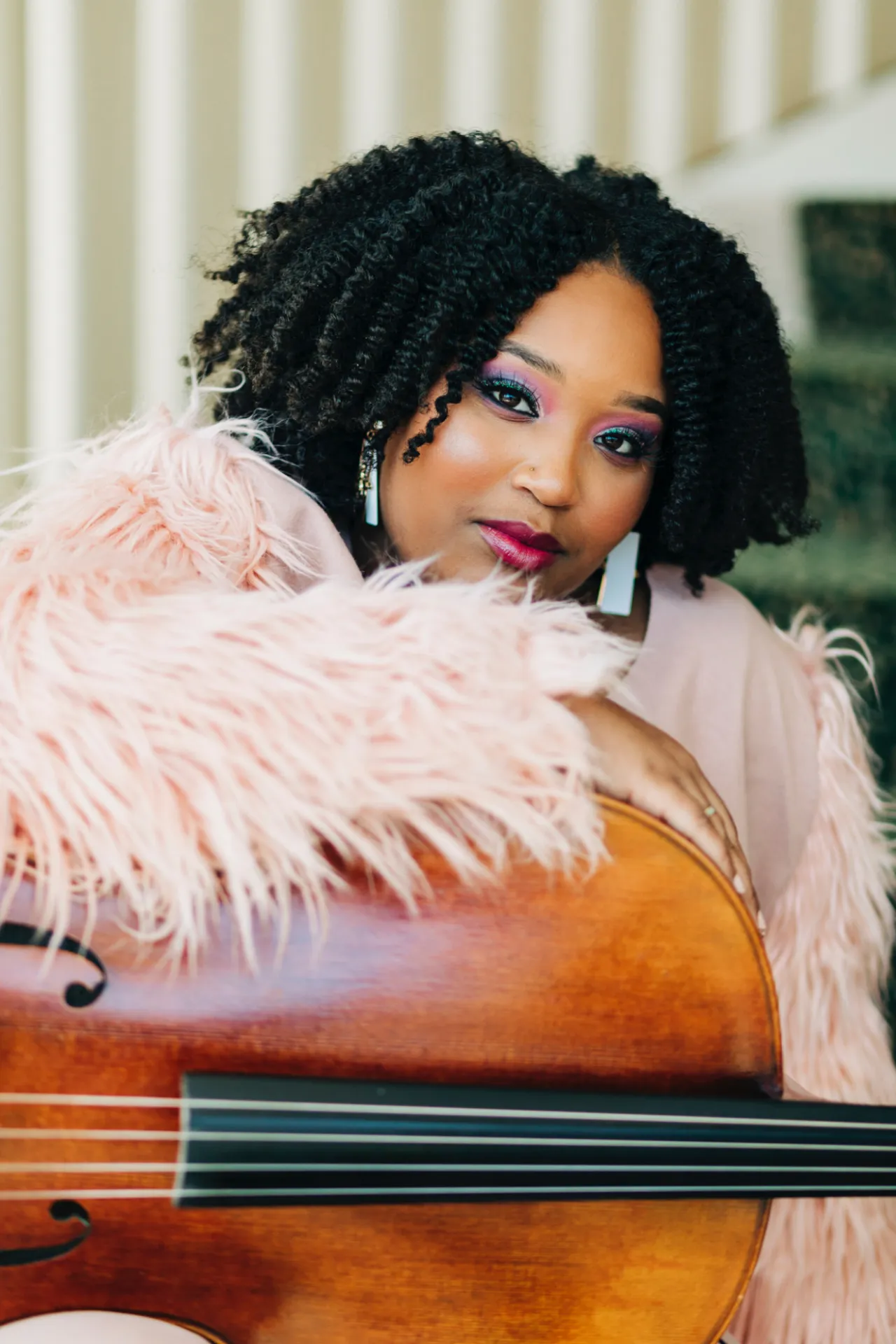 Sasha Doster wears a feathery pink top and holds her cello in her lap. She is sitting on a green and white staircase.