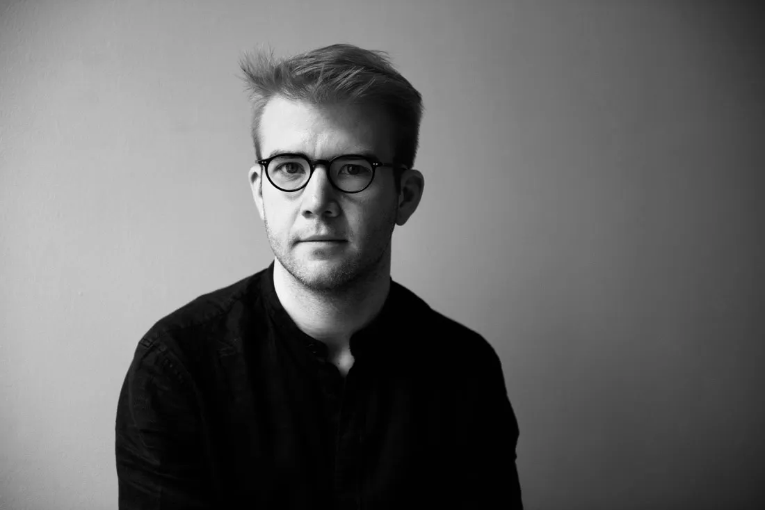 Parker Ramsay in black and white. He is wearing a black shirt against an ombre gray background. He has dark rimmed glasses.