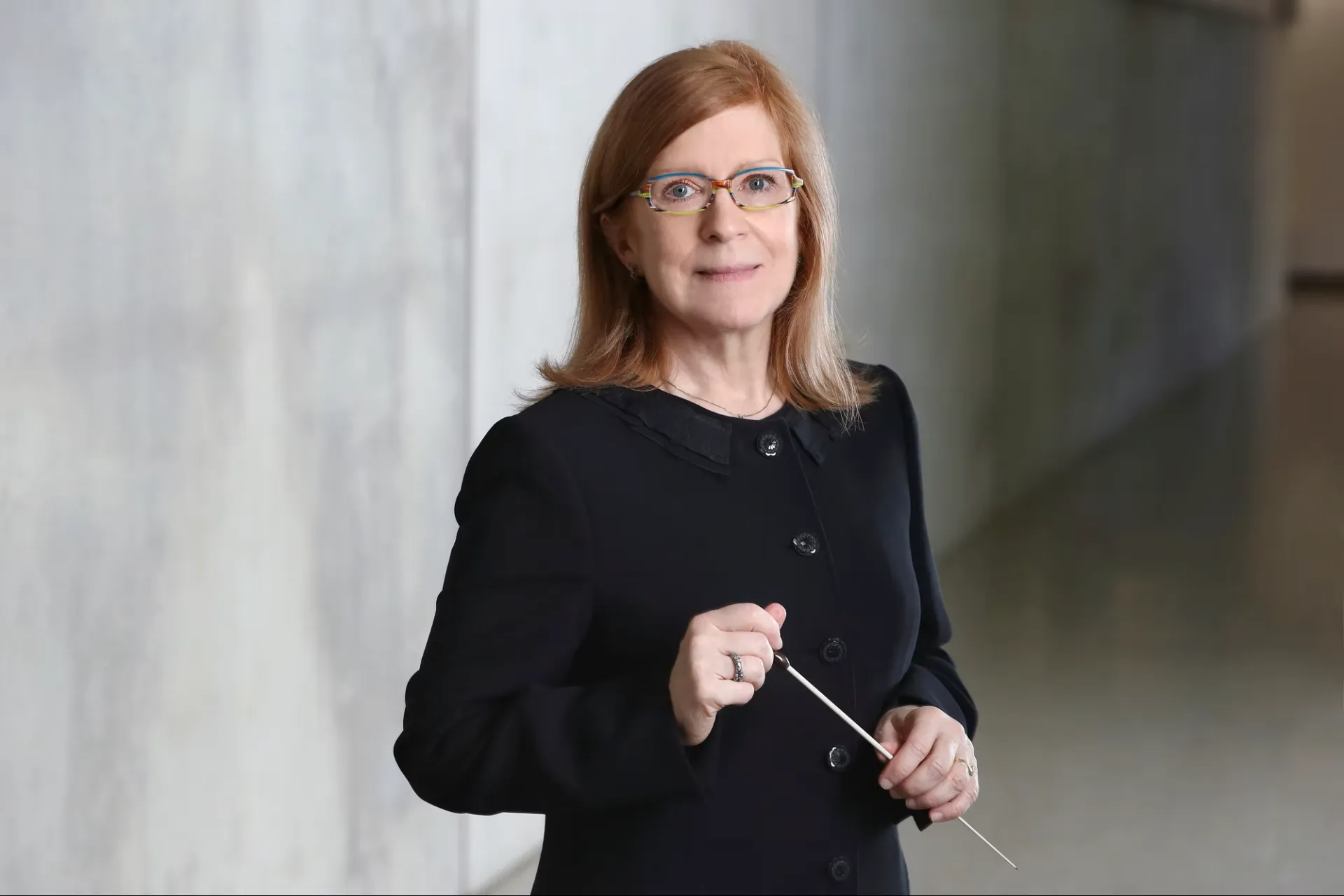 Emily Brown stands against a gray background. She has colorful glasses, red hair, and a black dress with buttons down the middle. She holds a white conductor's baton.