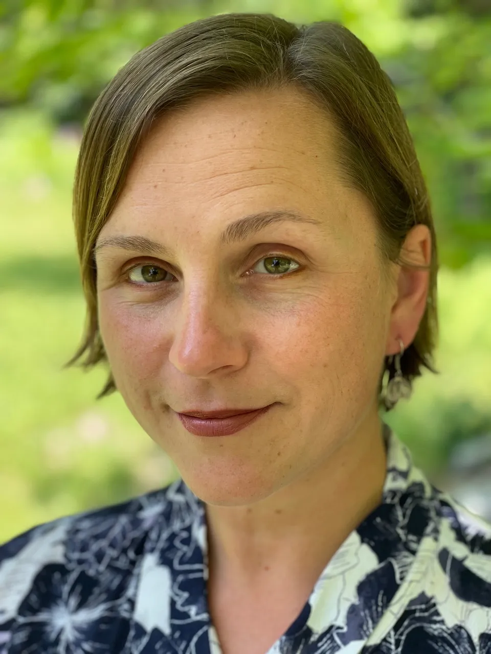 Maria Sonevytsky looks at the camera with a slight smile, greenery behind her, and a blue and white shirt.