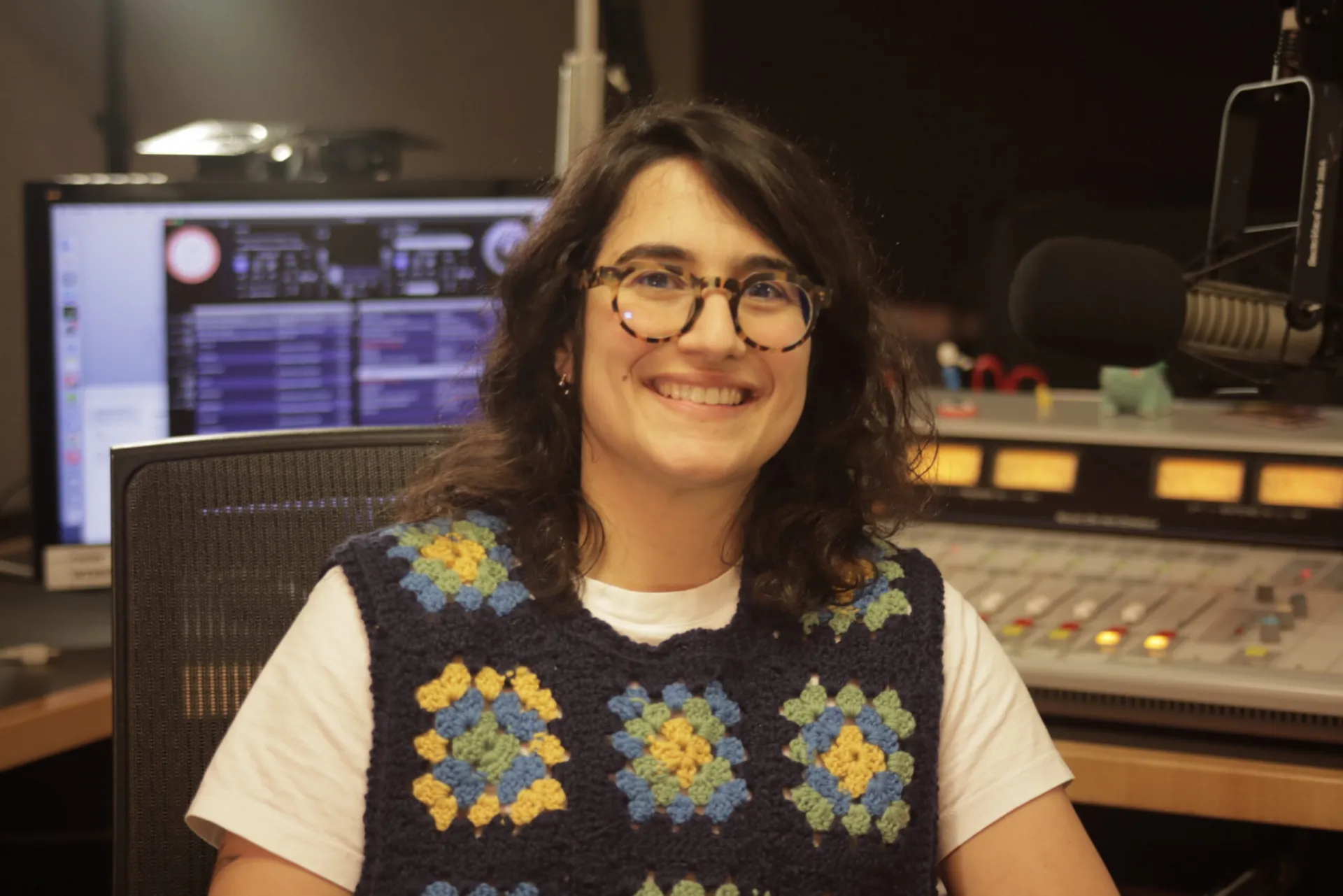 Paola Cossermelli Messina sits in a radio studio with a mixing board behind her. She's smiling at the camera in a knit vest and horn-rimmed glasses.