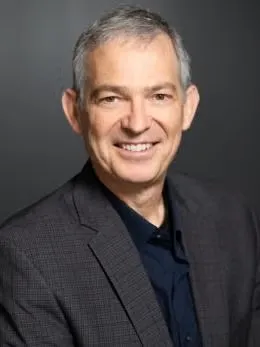 Tim Weiss smiling in front of a gray background. He is wearing a darker gray suit jacket with a dark blue shirt.